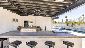 Un área de bar al aire libre con mostrador de concreto, cuatro taburetes de bar tejidos, un ventilador de techo y una piscina al fondo.