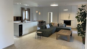 Ett modernt vardagsrum med en grå hörnsoffa, ett soffbord i trä och en tv. Det finns också en köksdel med ett matbord och stolar, samt en öppen spis med en växt bredvid.