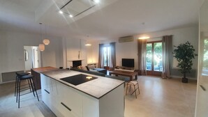 Ett modernt kök med en central köksö, barstolar, en matplats med bord och stolar, en tv och ett stort fönster med gardiner.