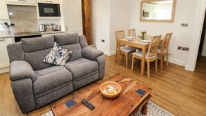 Un salon moderne avec un canapé gris, une table à manger en bois et un coin cuisine doté de meubles blancs et d’un four encastré.