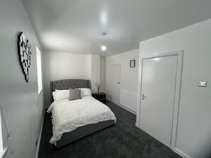 A modern bedroom with a grey upholstered bed, a white headboard, and a dark grey carpet. There is a white door on the right and a white radiator on the wall. - Modern Executive 2-bed Apartment in London (London)