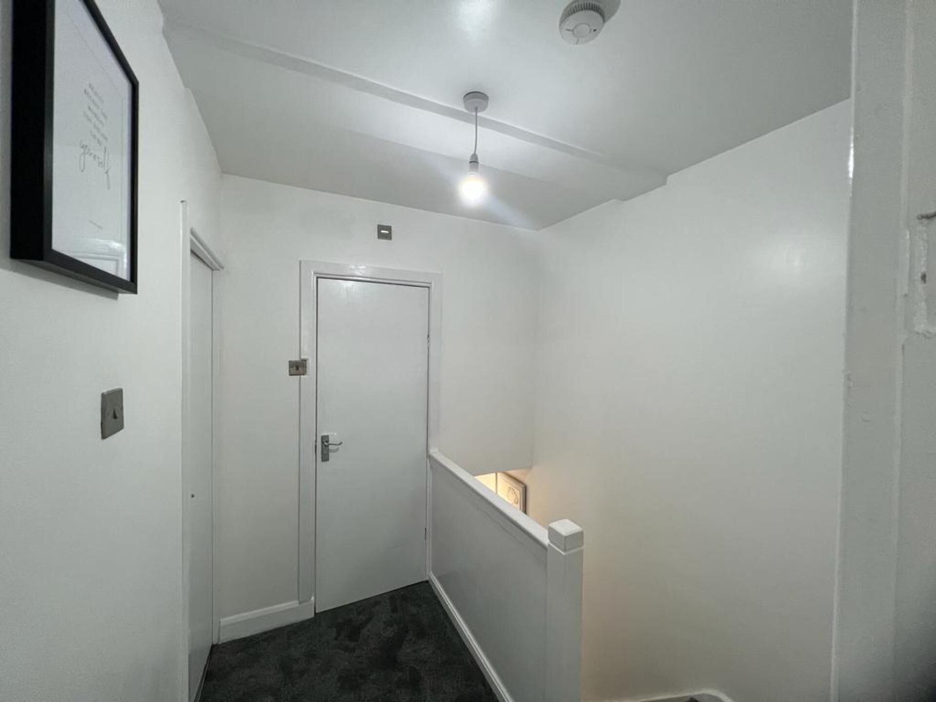 Un passadís estret amb una porta blanca, una llum al sostre i un quadre a la paret.