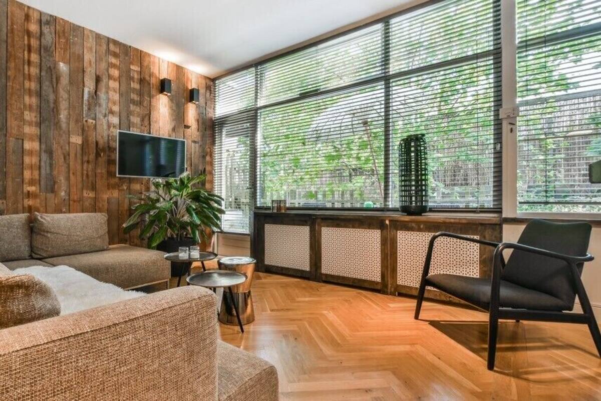 Uma sala de estar moderna com painel de parede de madeira, uma janela grande com persianas, um sofá, uma poltrona e uma mesa pequena.