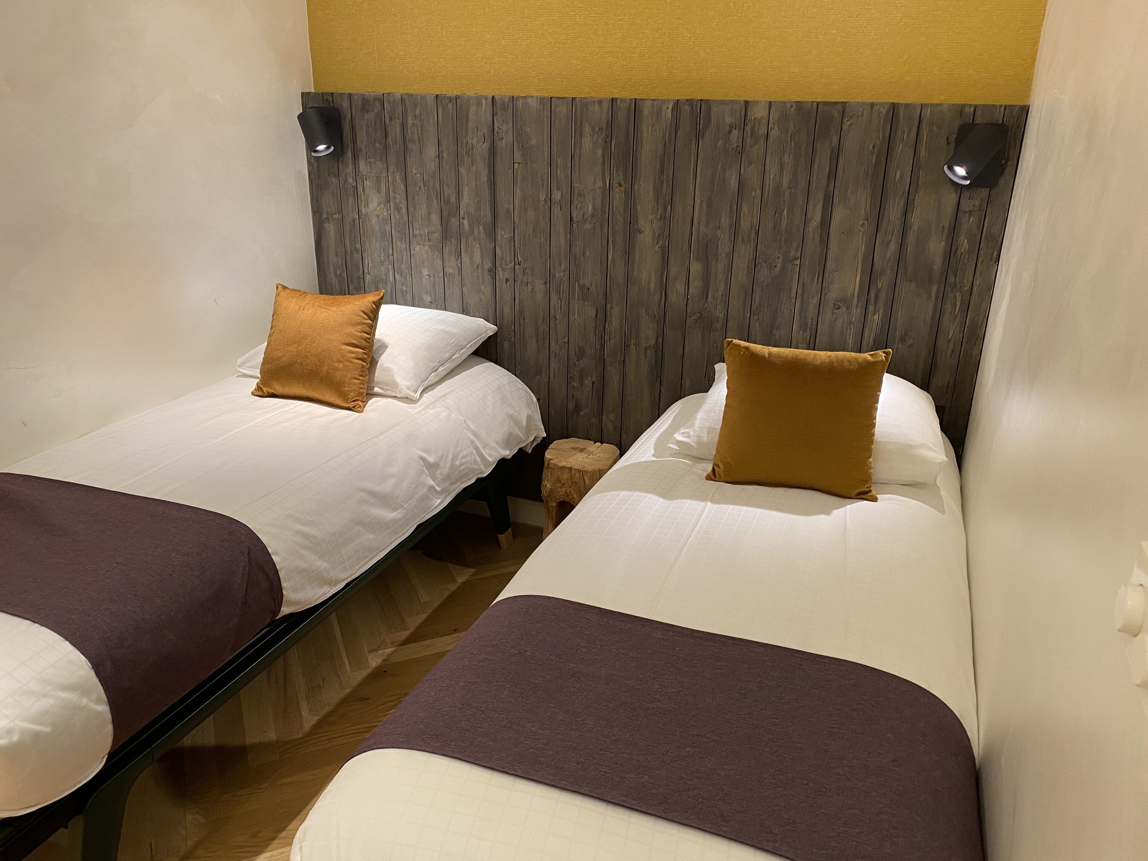 Habitación de hotel con dos camas individuales, cada una con almohada marrón y ropa de cama blanca. La habitación tiene un cabecero de madera y un taburete de madera pequeño.