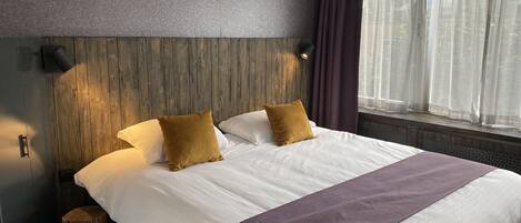 A neatly made bed with white linens and two mustard pillows, a wooden headboard, a wall-mounted air conditioner, and a window with sheer curtains.