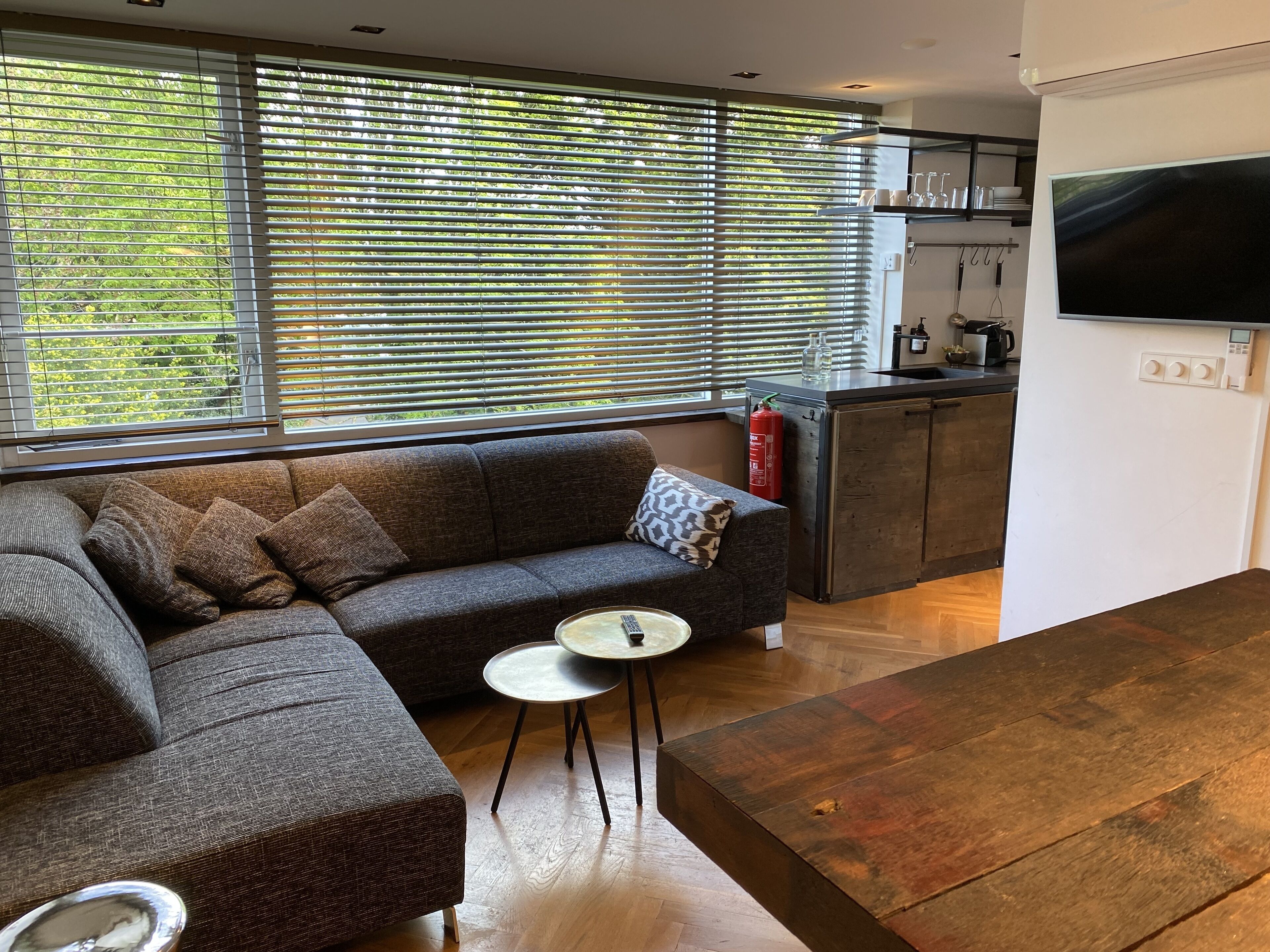 A modern living room with a sectional sofa, a small round table, and a wooden coffee table. There is a kitchen area with a built-in oven and a large flat-screen TV mounted on the wall.