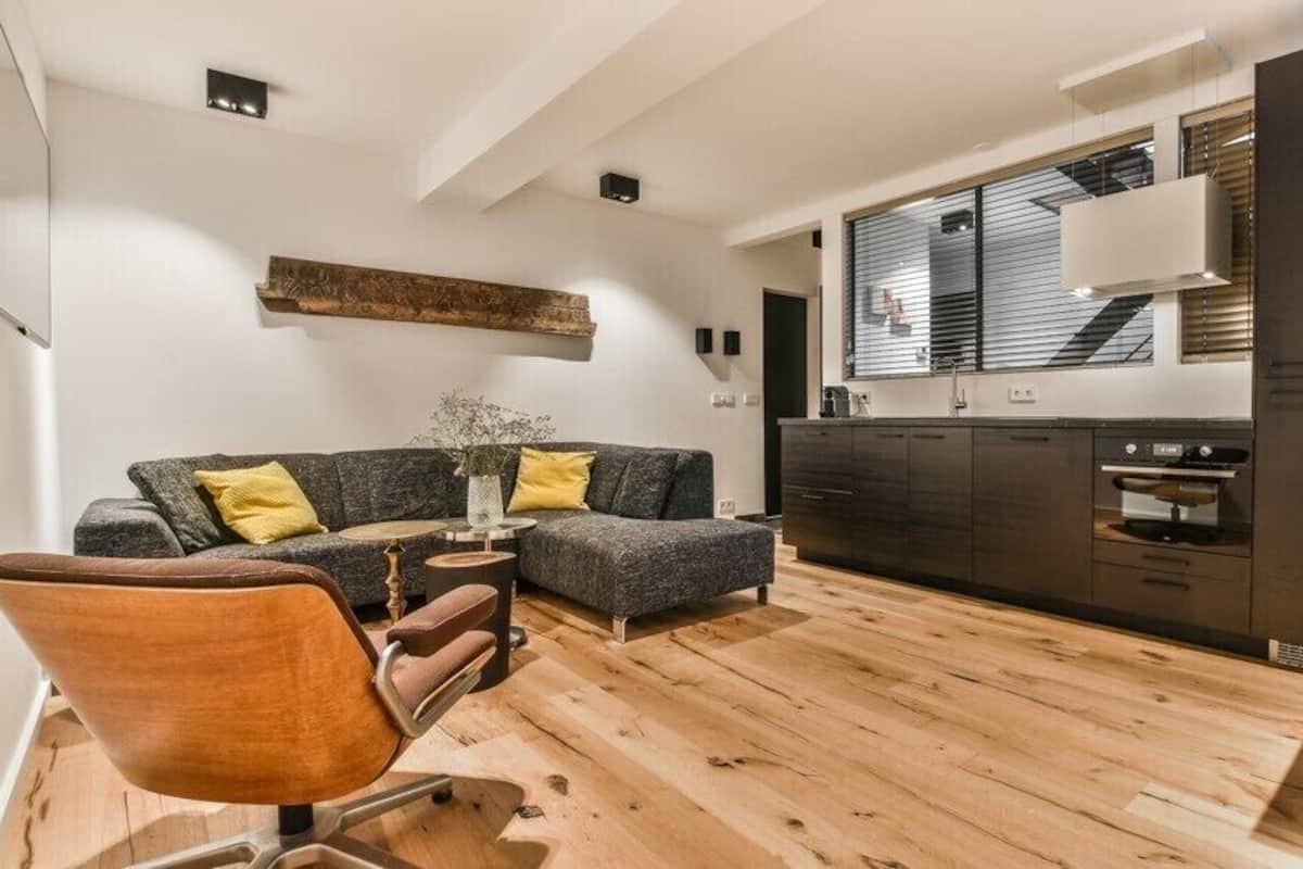 Uma sala moderna com sofá, poltrona e mesa de centro. Cozinha com armários escuros e uma janela é visível ao fundo.