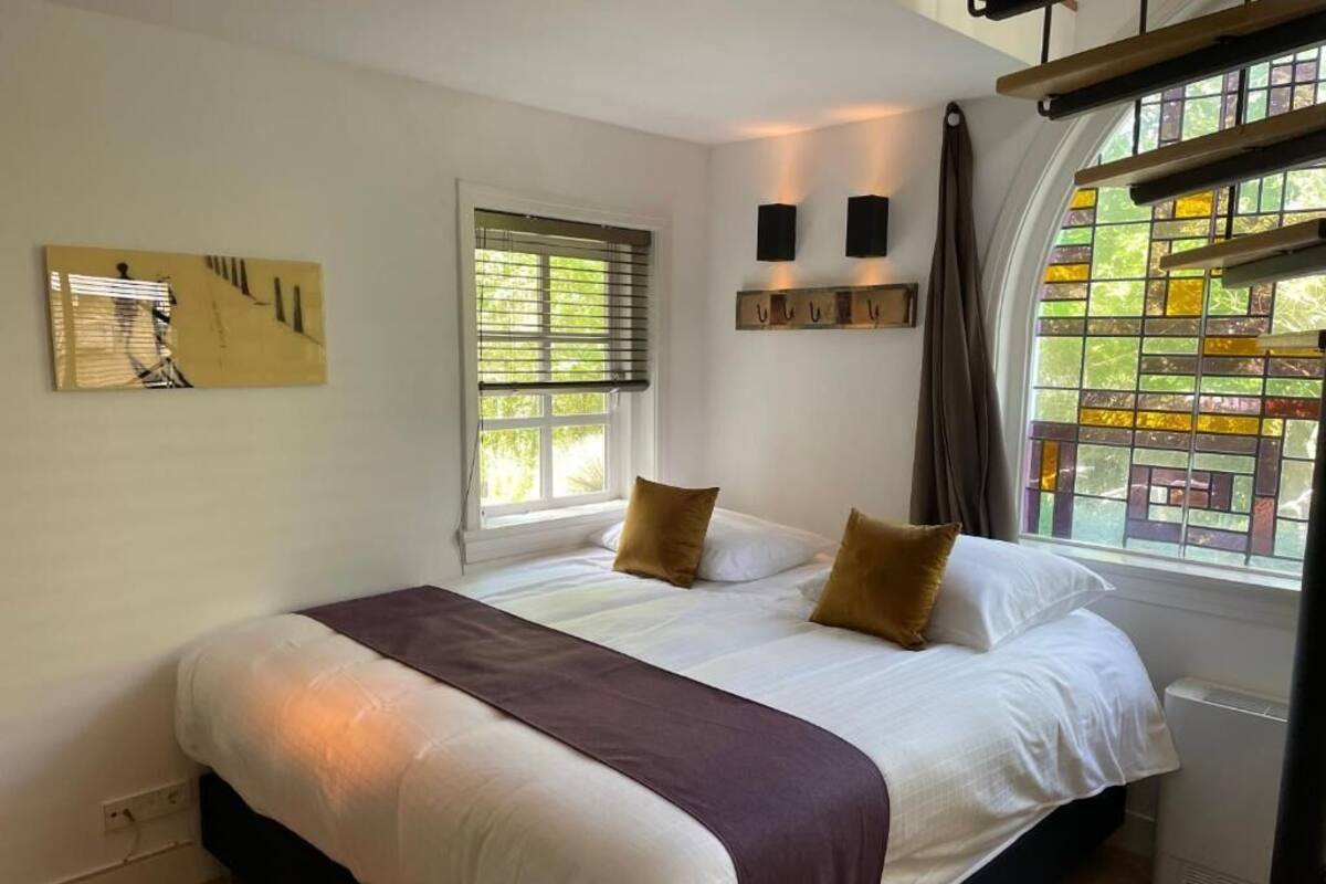 Quarto com cama grande, duas almofadas, mesa de cabeceira, luminária na parede, janela com persianas e vista para o verde lá fora.