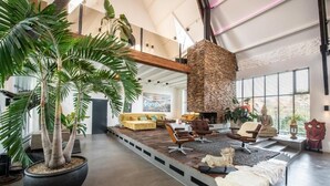 A spacious living room with a large potted plant, a stone fireplace, and a high ceiling with exposed beams. - het Lichthuis (Zandvoort)