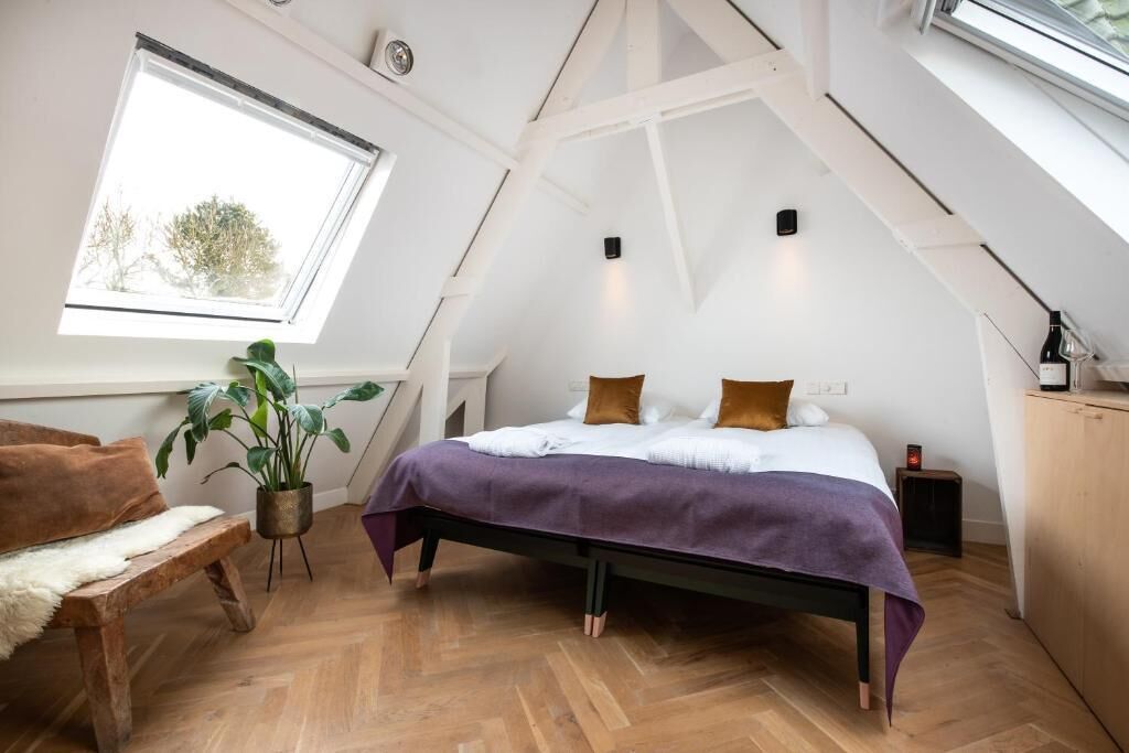 A bedroom with a sloped ceiling, a bed with a purple bedspread, a wooden bench, a potted plant, and a window with a view of trees.