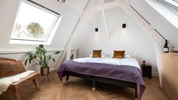 Ein Schlafzimmer mit geneigter Decke, einem Bett mit violetter Bettdecke, einer hölzernen Bank, einer Topfpflanze und einem Fenster mit Blick auf Bäume.