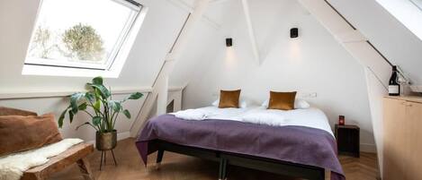 A bedroom with a sloped ceiling, a bed with a purple bedspread, a wooden bench, a potted plant, and a window with a view of trees.