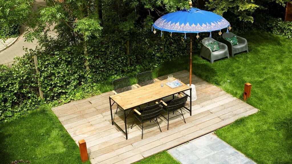 Un terrazzo in legno con tavolo e sedie, immerso nel verde e ombreggiato da un ombrellone blu.