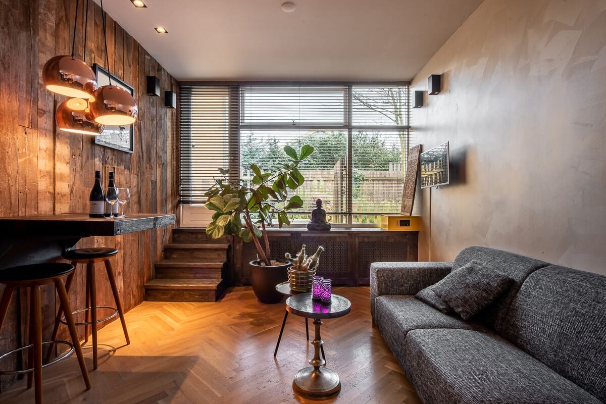 Uma sala de estar moderna com parede de madeira, sofá cinzento, mesa de centro redonda e um espaço bar com banquetas.