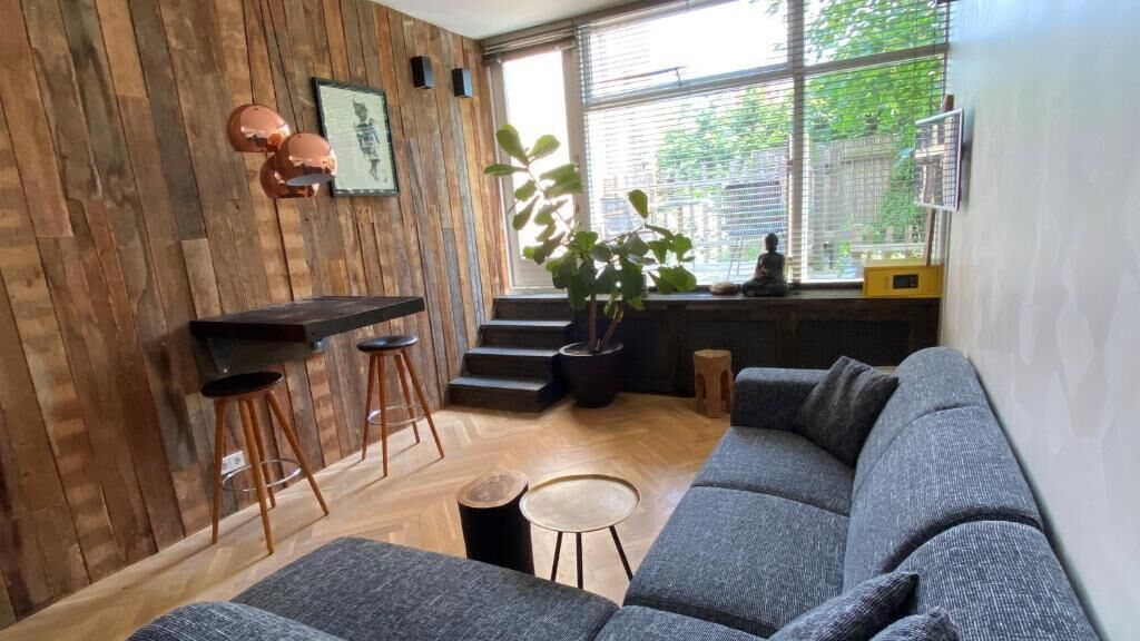 Um espaço de estar moderno com parede de madeira, sofá cinza, um pequeno bar com banquetas e um vaso de planta.