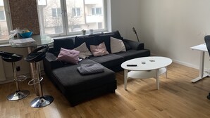 A modern living room with a black sectional sofa, a glass-top coffee table, and a white side table. A black guitar leans against the wall. - Charming Apartmemt in Stockholm City (Stockholm)