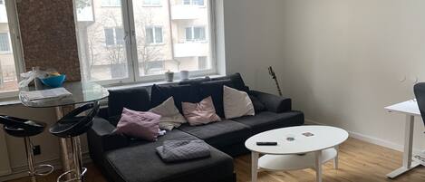 A modern living room with a black sectional sofa, a glass-top coffee table, and a white side table. A black guitar leans against the wall.