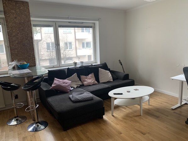 A modern living room with a black sectional sofa, a glass-top coffee table, and a white side table. A black guitar leans against the wall. - Charming Apartmemt in Stockholm City (Stockholm)