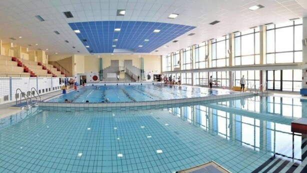 En inomhuspool med flera banor, en central trappa och ett område med sittplatser och läktare.