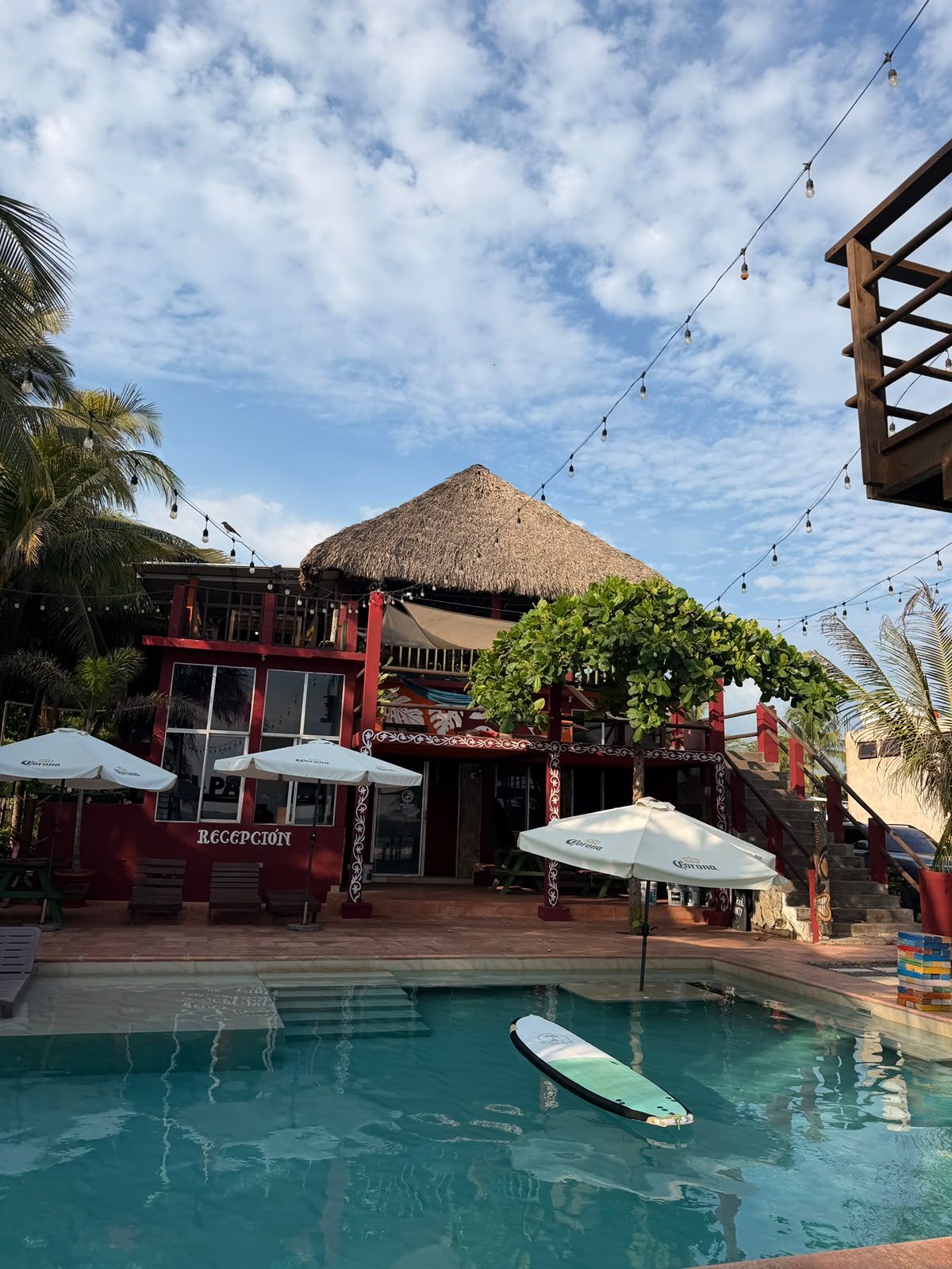 Zona de piscina con una estructura de techo de paja, sombrillas y un cartel de recepción.