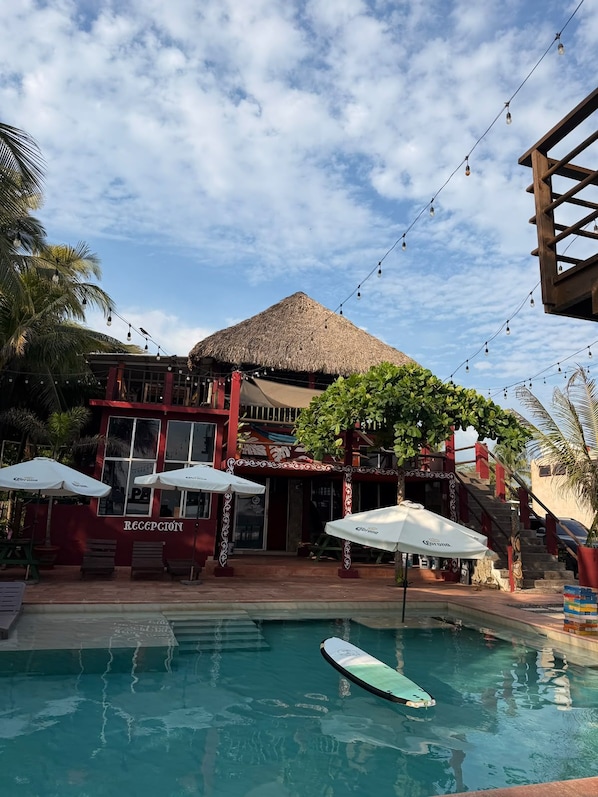 A pool area with a thatched-roof structure, umbrellas, and a reception sign.