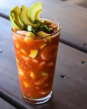 Un vaso alto con una bebida roja, posiblemente un cóctel, adornado con rodajas de aguacate y hierbas.