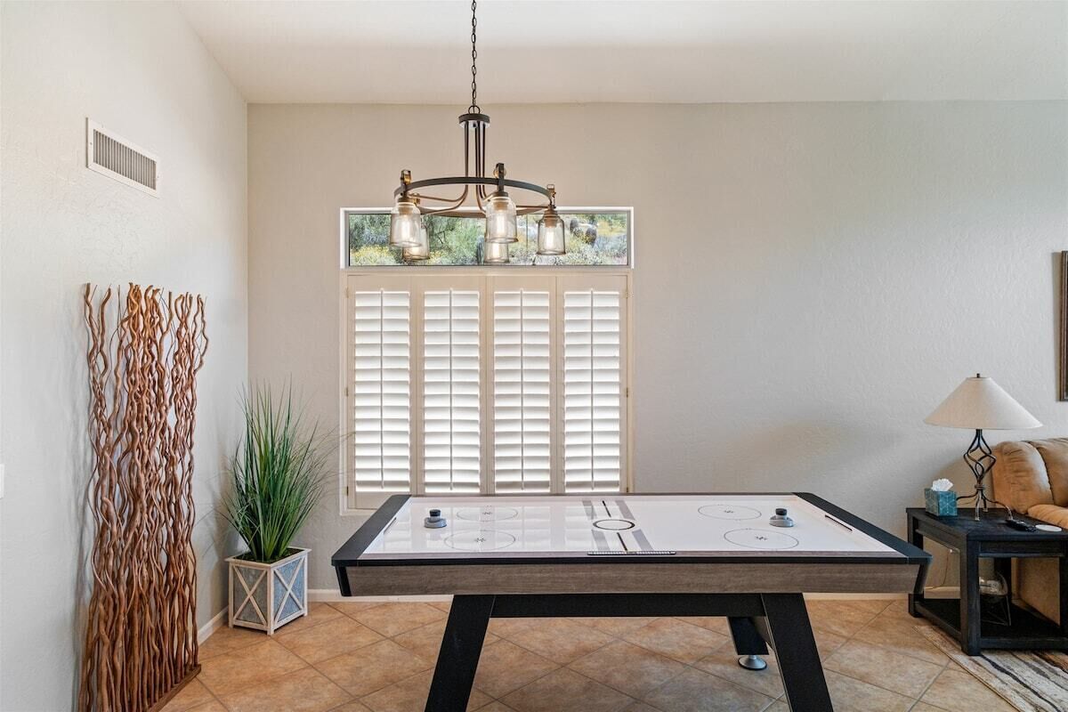 Une pièce avec une table de billard, un lustre, une fenêtre avec des stores et une plante.