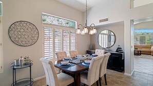 A dining area with a wooden table, beige chairs, and a chandelier. There is a window with blinds, a round mirror, and a black cabinet.