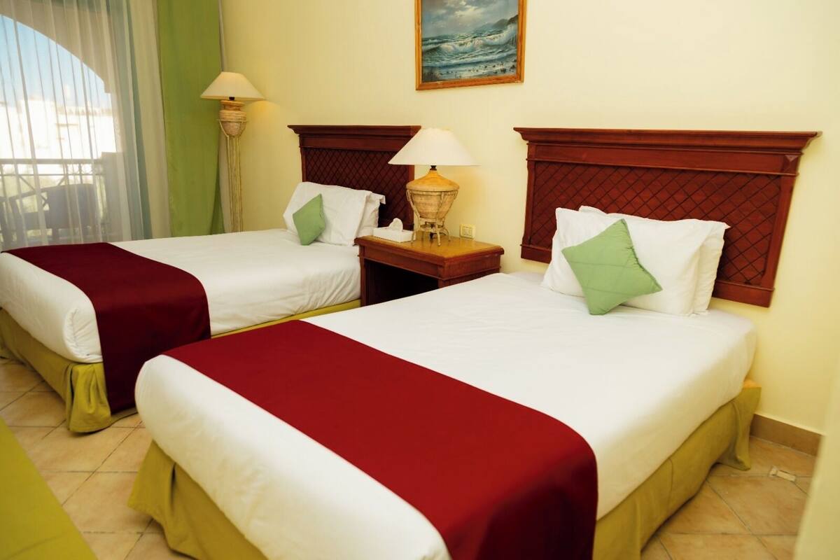 Habitación de hotel con dos camas, cada una con cubrecama rojo y blanco, almohadas verdes y cabeceros de madera. Hay una mesita de noche con una lámpara entre las camas, y un cuadro de un paisaje marino en la pared.