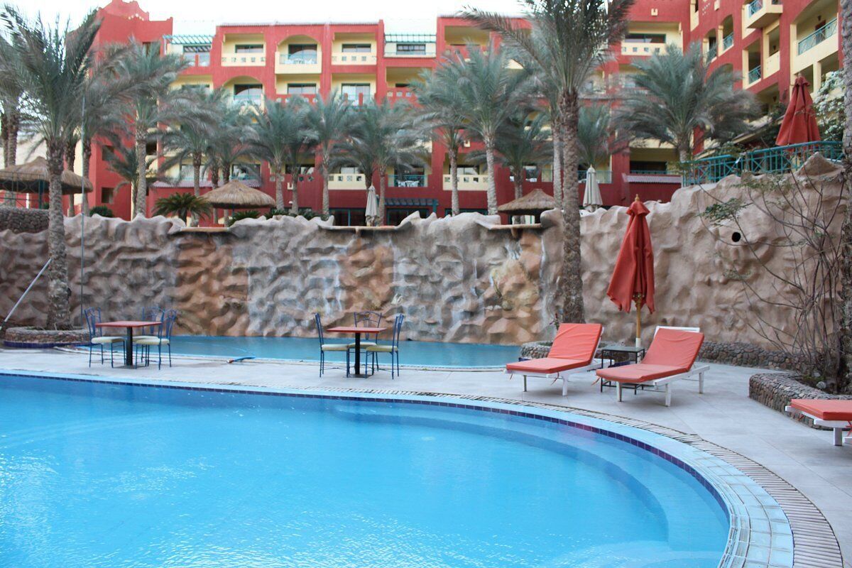 A swimming pool with lounge chairs and tables, surrounded by palm trees and a rocky wall.