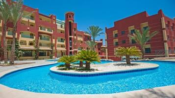 A hotel with a swimming pool surrounded by palm trees and a red building facade.