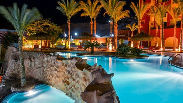 Une piscine nocturne, avec une formation rocheuse, des palmiers et un bâtiment éclairé de lumières chaleureuses en arrière-plan.