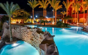 A nighttime swimming pool with a rock formation, palm trees, and a building with warm lighting in the background. - Sun & Sea Hotel and Aqua Park - Hurghada (Hurghada)