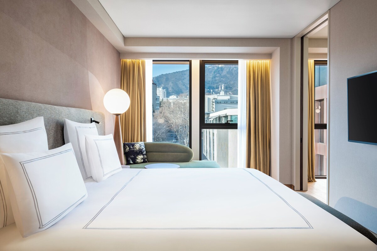A hotel room with a large bed, a view of a cityscape, and a flat-screen TV.