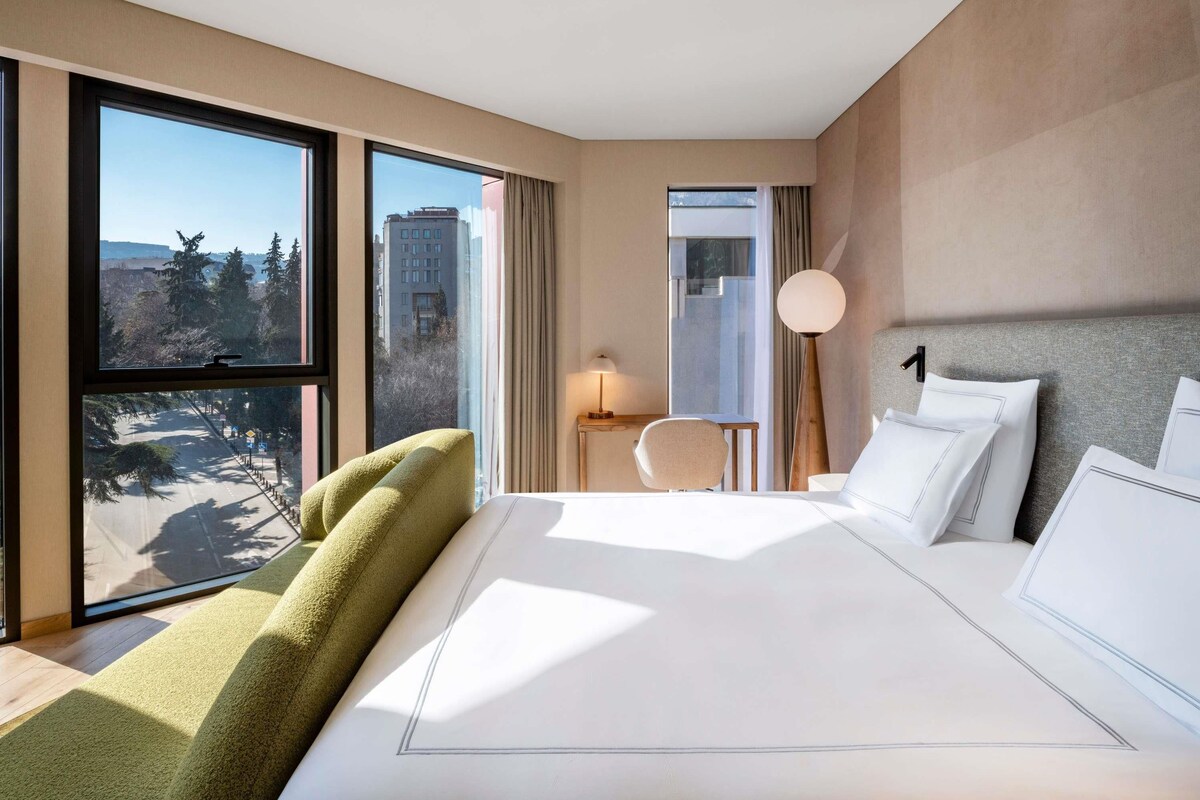 A modern hotel room with a large bed, a view of the city, and a desk with a lamp.