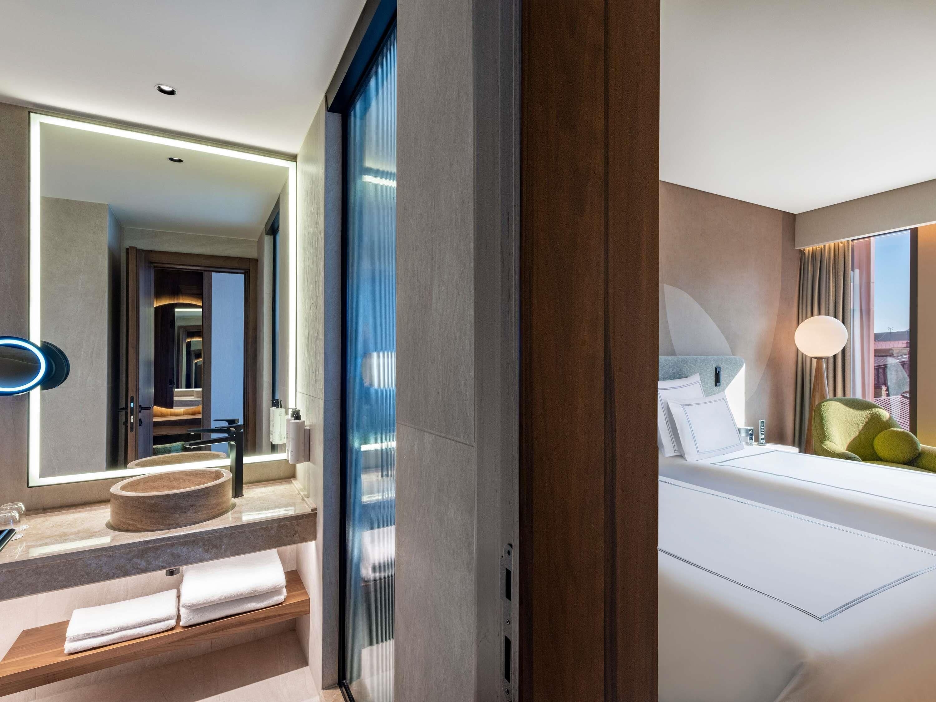 A modern hotel room with a large bed, a bedside table, a mirror, and a view of the city.