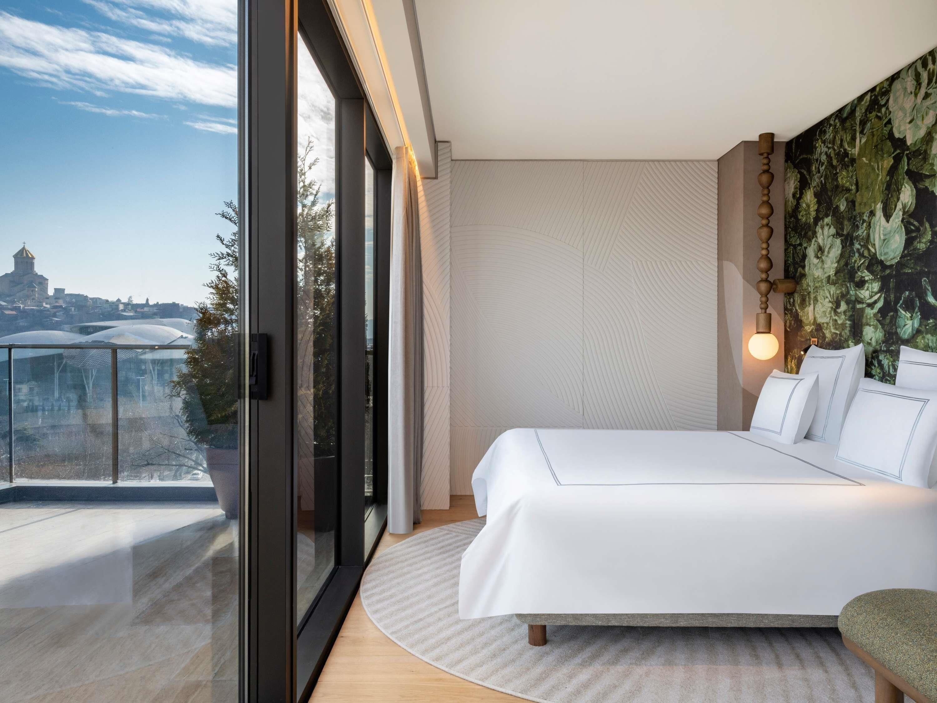 A modern bedroom with a large bed, a view of a cityscape, and a balcony with glass doors.