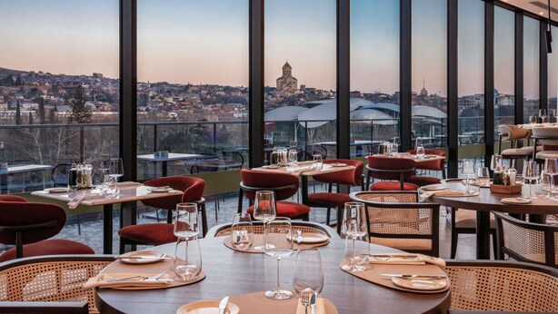 A restaurant with tables set for dining, large windows offering a city view, and a modern interior design. - Swissôtel Tbilisi (Tbilisi)