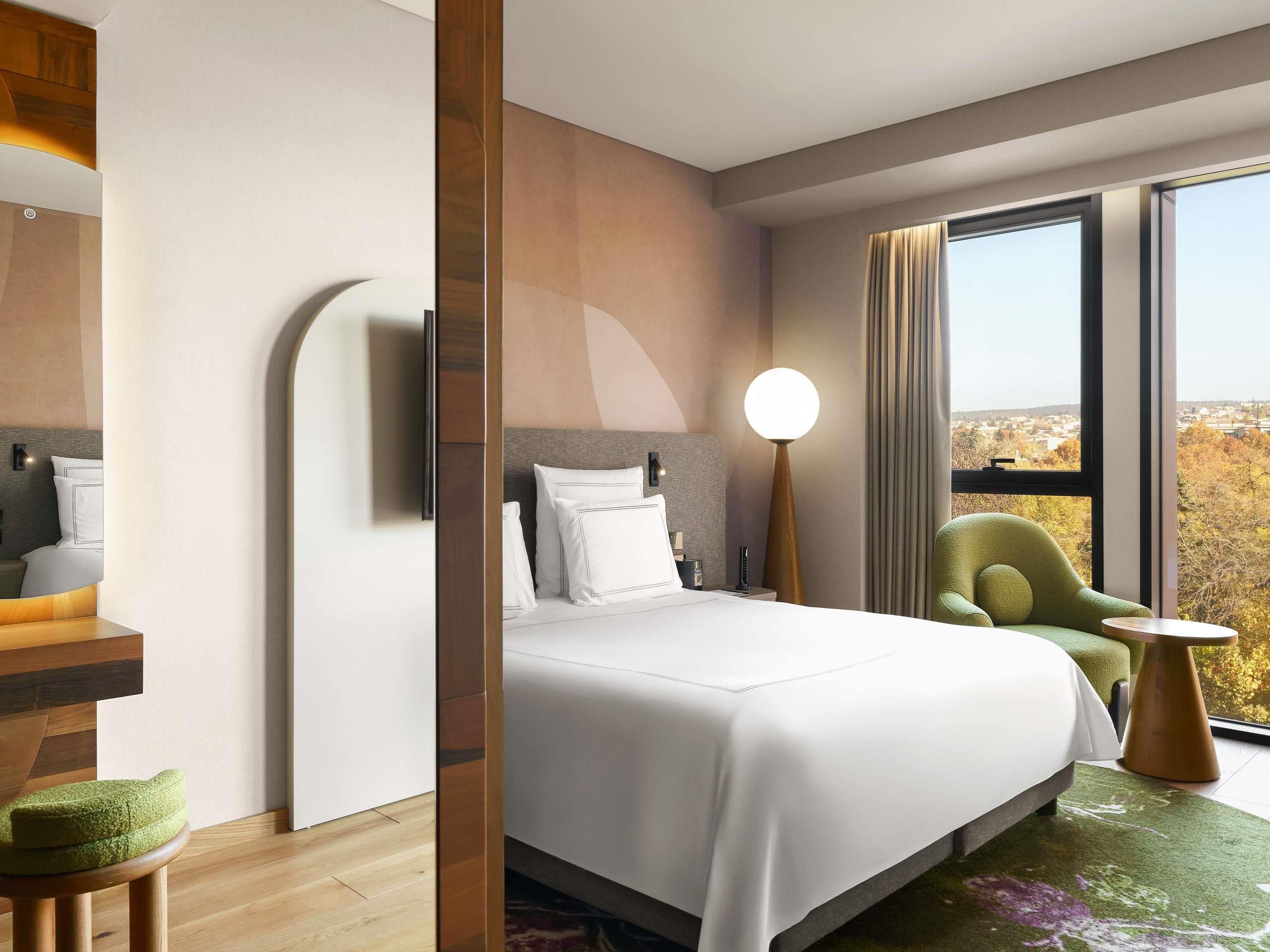 Une chambre d’hôtel moderne avec un lit, un fauteuil vert, une table ronde et une vue sur la ville.