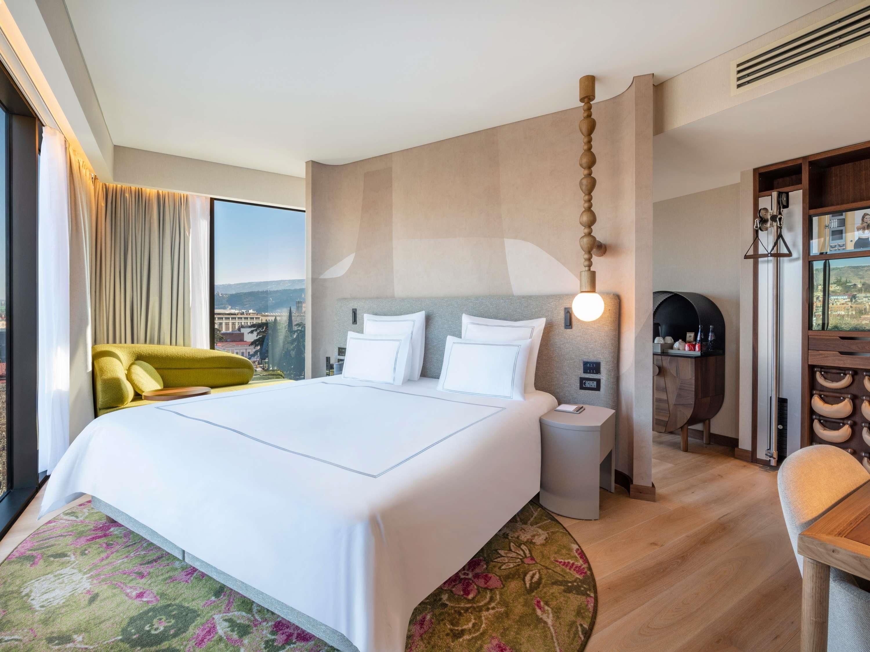 A hotel room with a large bed, a nightstand, a chair, a desk, and a view of the city.