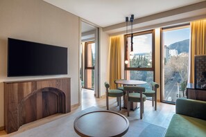 A modern hotel room with a flat-screen TV, a wooden fireplace, a round dining table with chairs, and large windows offering a city view. - Swissôtel Tbilisi (Tbilisi)