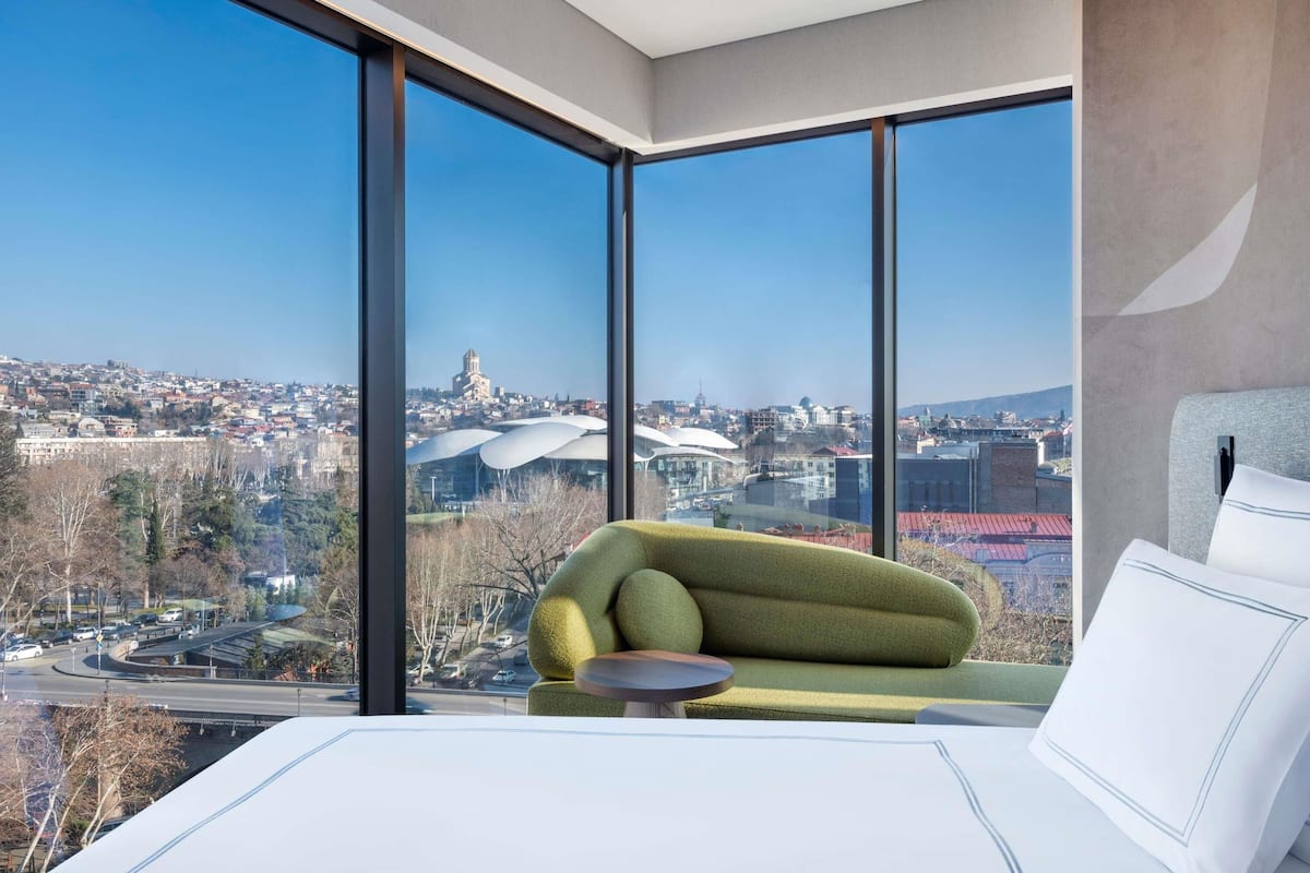 A modern hotel room with a large window offering a city view, a green sofa, and a bed with white bedding.
