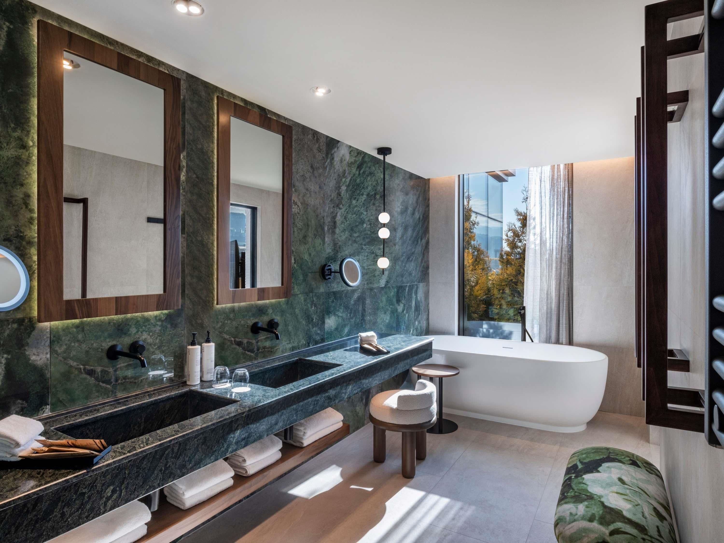 A modern bathroom with a dual sink vanity, a large mirror, a round mirror, a bathtub, and a view of trees through the window.