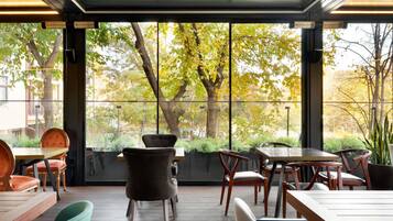Un área de estar interior con mesas y sillas, ventanas amplias que ofrecen vistas a árboles y un edificio.
