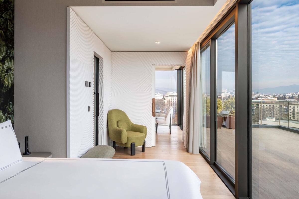 A modern bedroom with a large bed, a green armchair, and a balcony with a city view.