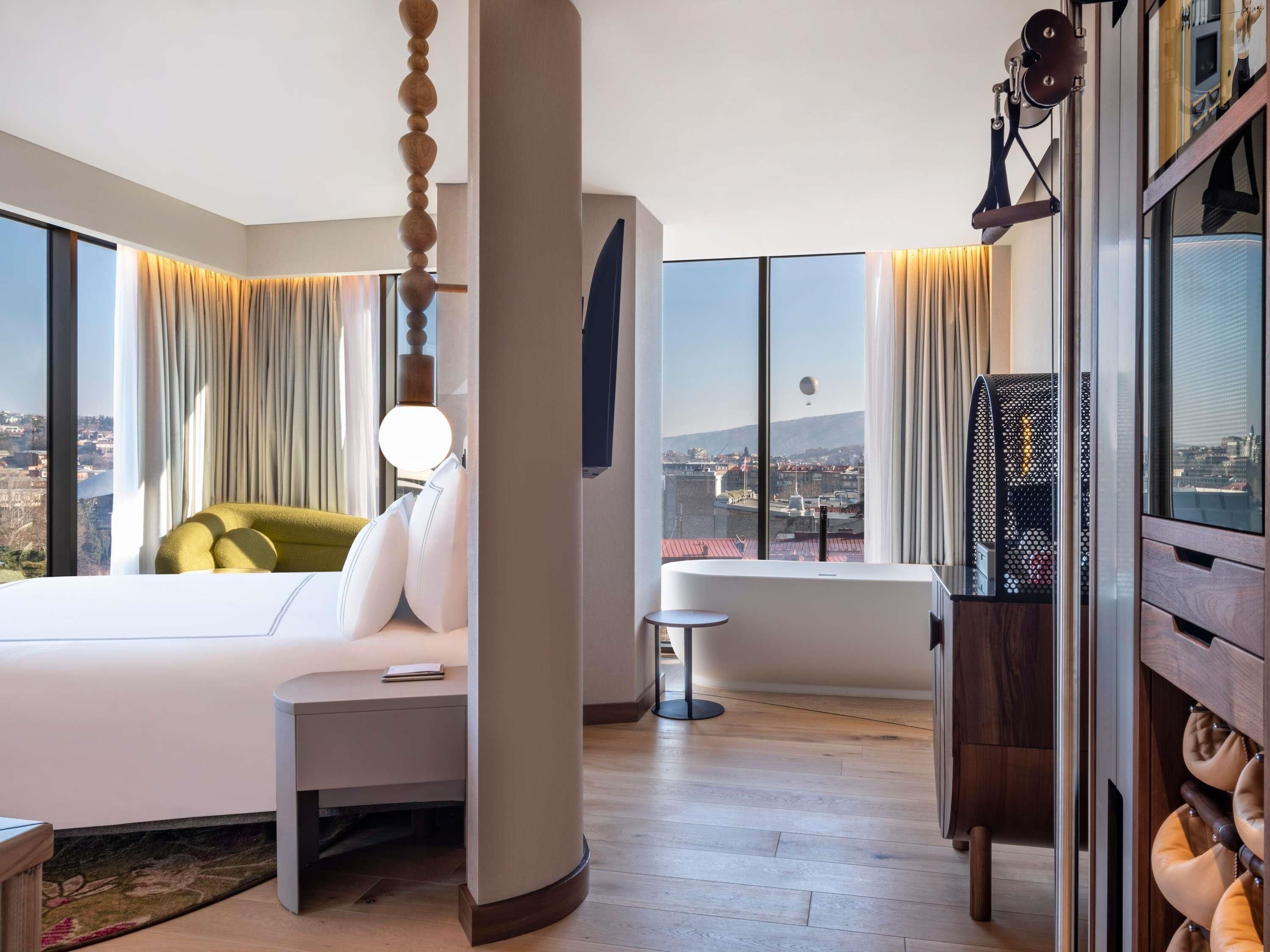 A modern hotel room with a large bed, a sitting area, a bathtub, and a city view.