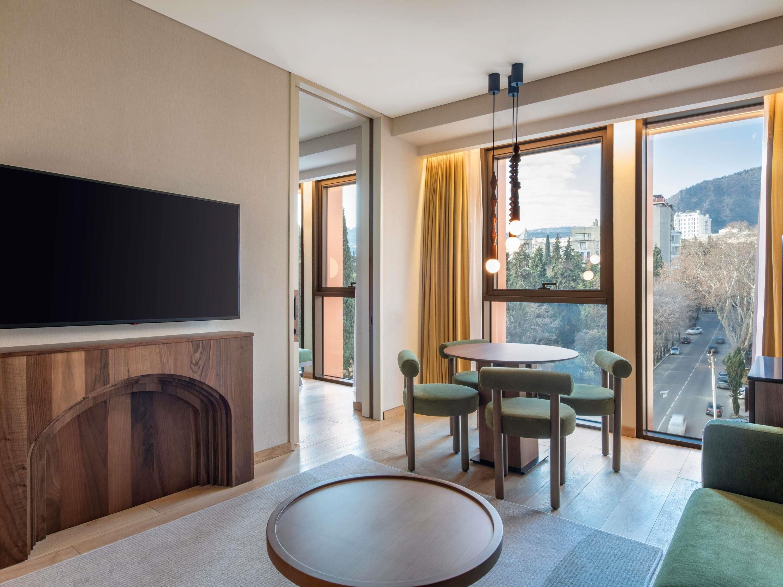 A modern hotel room with a flat-screen TV, a wooden cabinet, a round table, four green chairs, and large windows offering a city view.