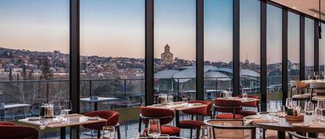 A restaurant with tables set for dining, large windows offering a city view, and a modern interior design.