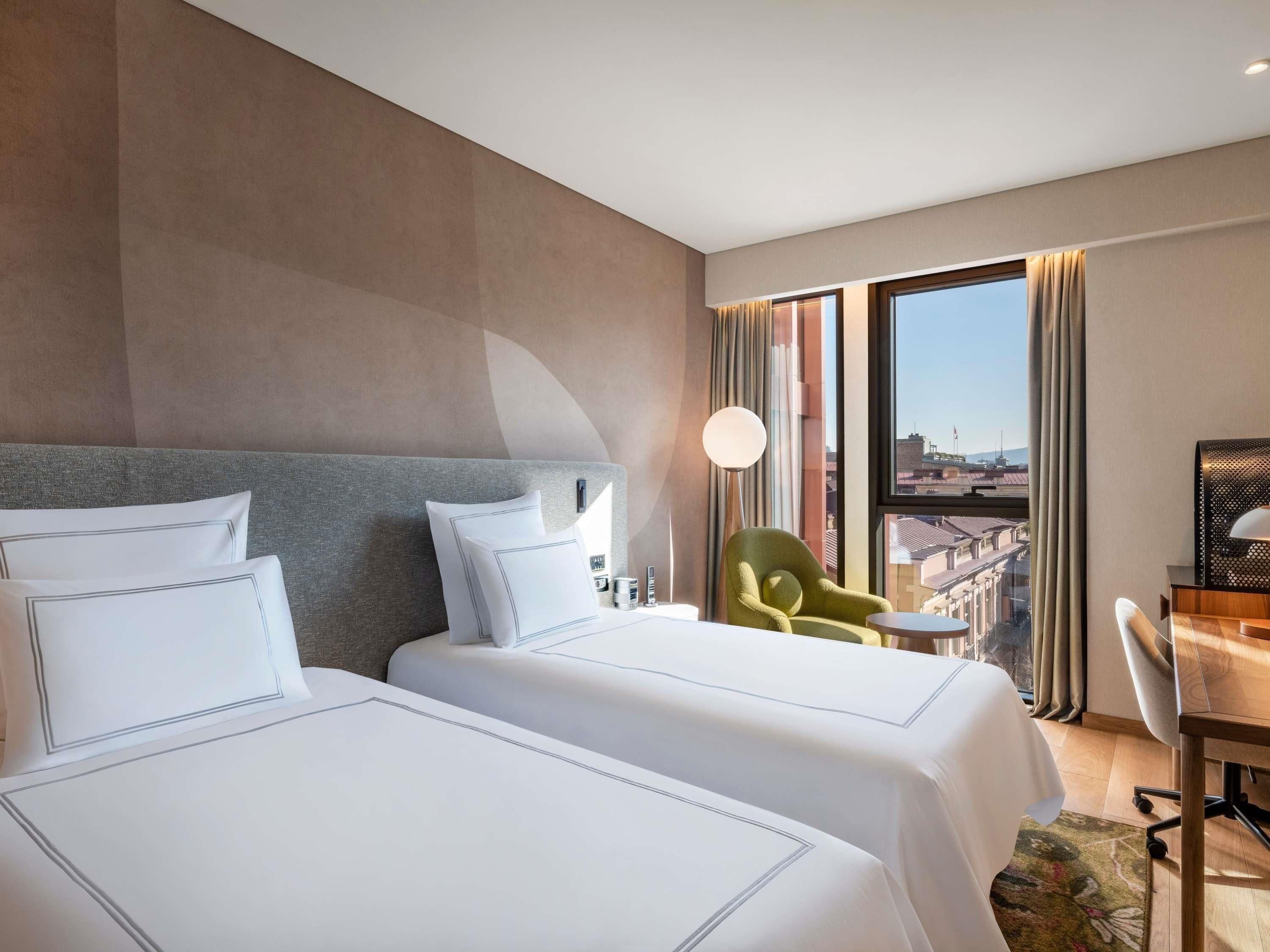 A hotel room with two beds, a desk, a chair, and a large window with a city view.