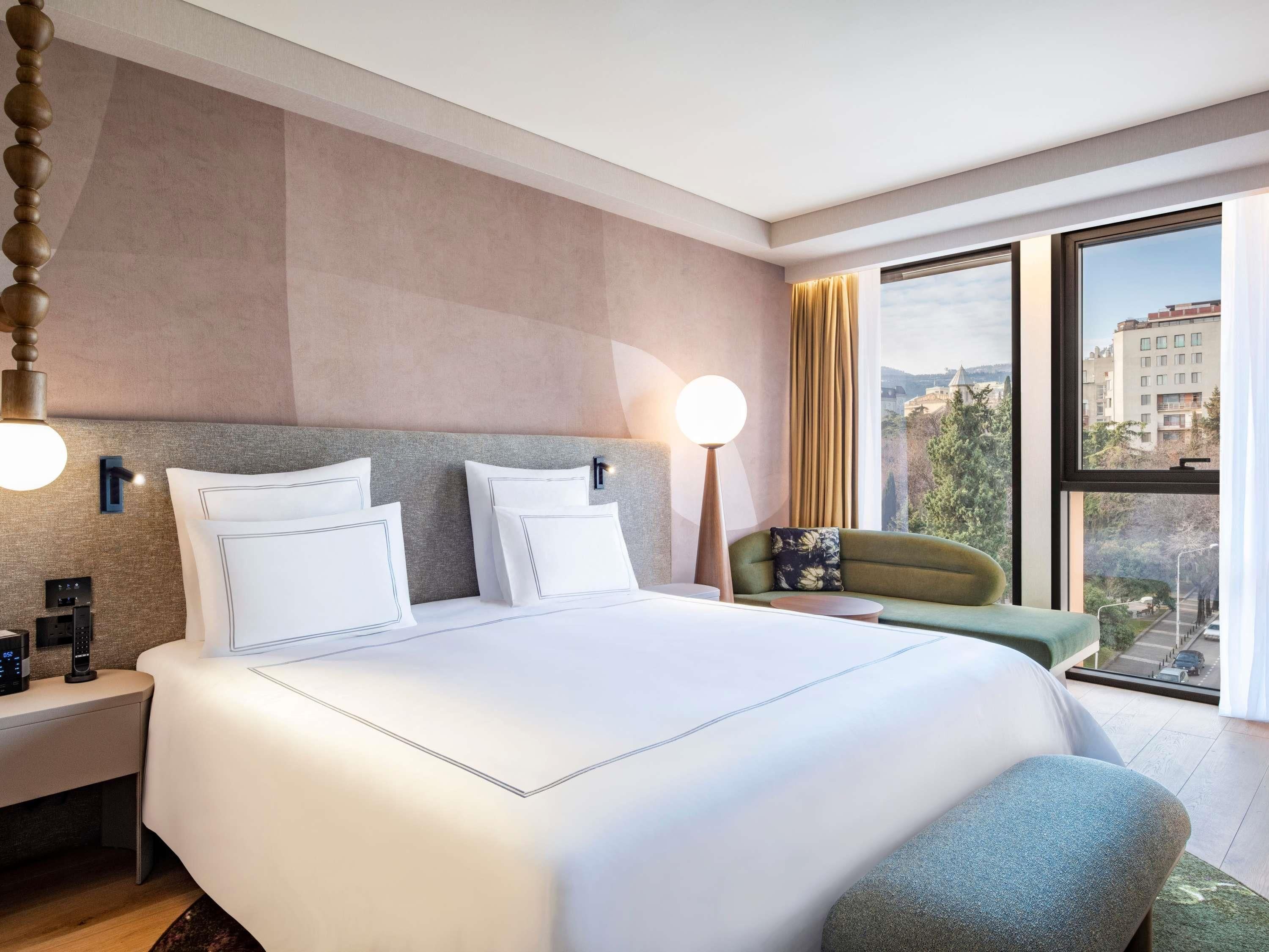 A hotel room with a large bed, bedside tables, a nightstand, a lamp, a chair, and a view of the city through large windows.